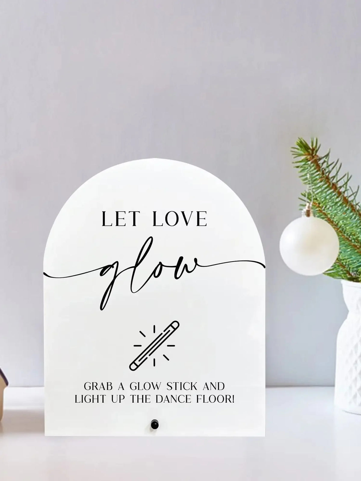 A white acrylic sign with black script that reads Let Love Glow and instructs to grab a glow stick for a dance floor.