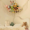 A multi-use storage hammock made of macrame is hanging on a wall, filled with various stuffed animals and plush toys.