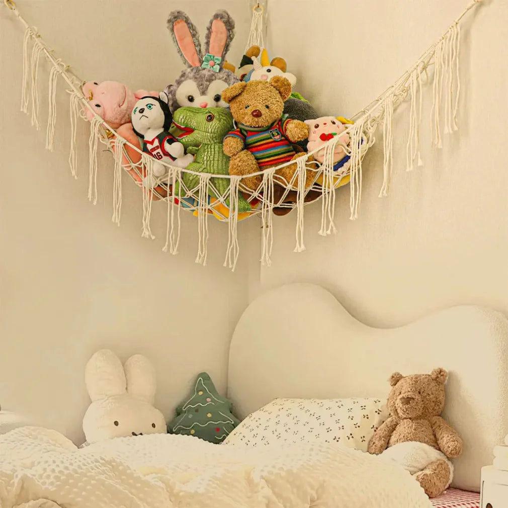 A multi-use storage hammock made of macrame is hanging on a wall, filled with various stuffed animals and plush toys.