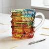 A stack of hand-painted book design mugs in vibrant shades of blue, green, and orange sitting on a white surface.