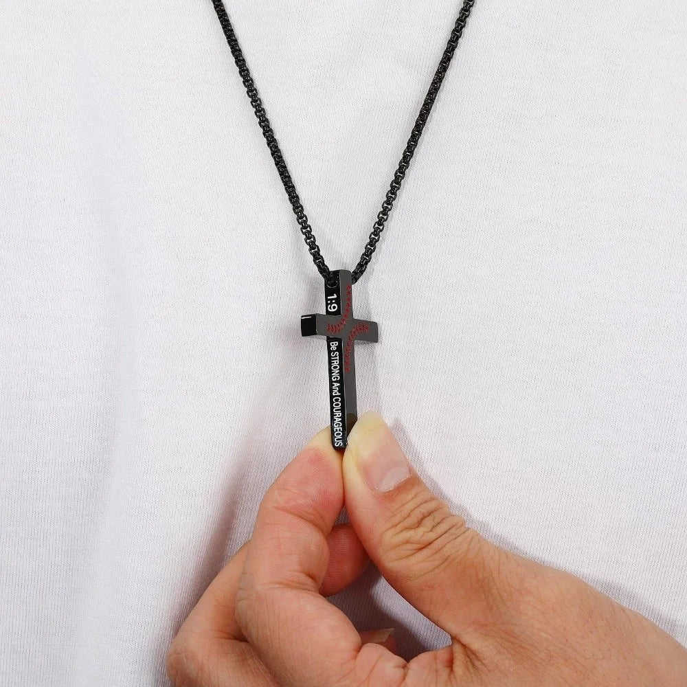 A black stainless steel cross pendant necklace with the inscription I'm strong and courageous is being held by a hand against
