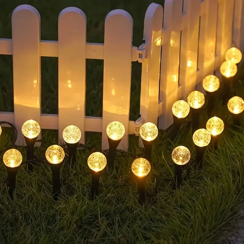 A row of white picket fence posts illuminates a lawn with glowing solar powered bubble ball garden lights.