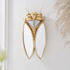 A golden cicada hanging ornament made of resin with iridescent wings mounted on a white wall.