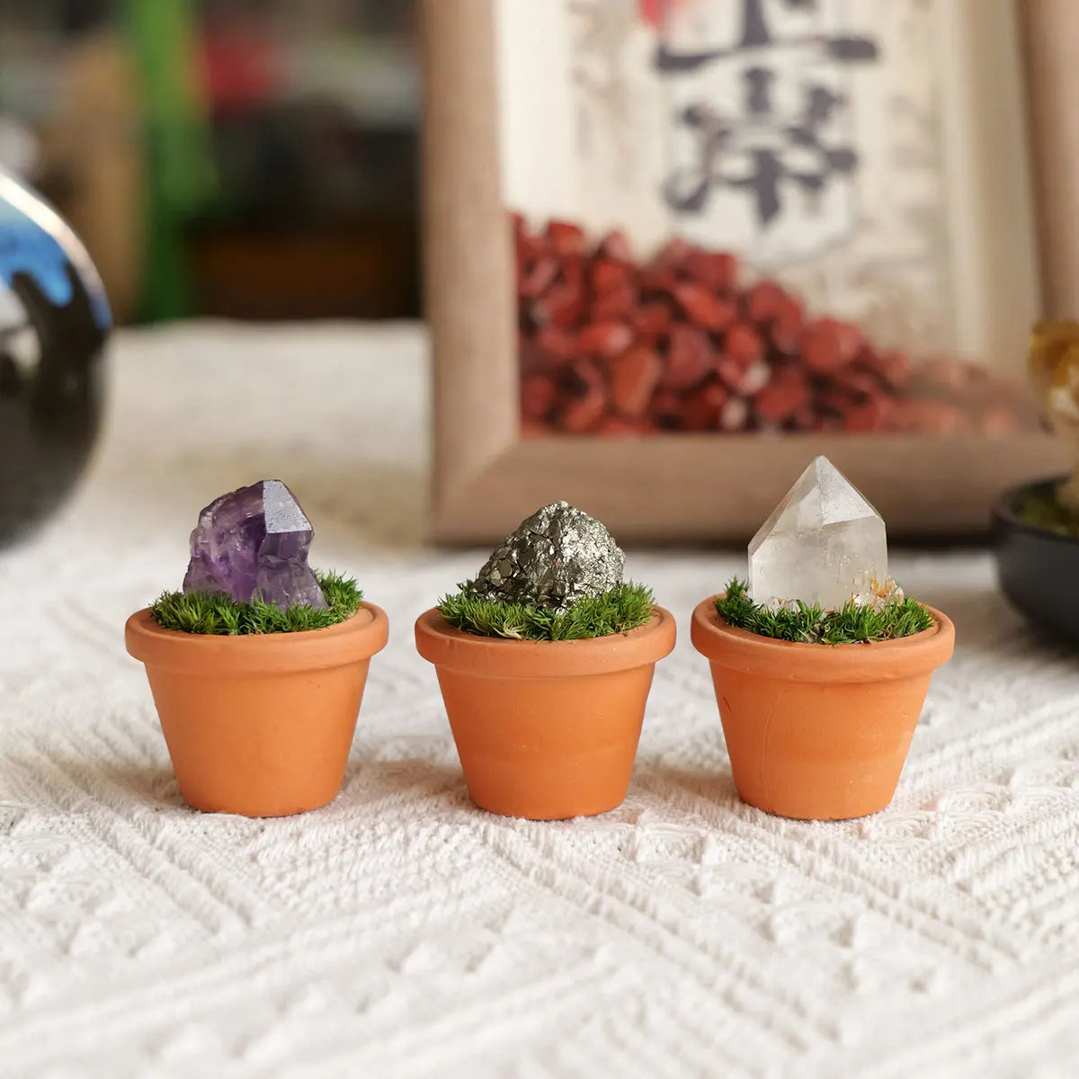 Three small terracotta pots hold amethyst and quartz crystals with green moss decoration.