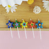 A row of colorful star-shaped birthday cake candles in blue, green, yellow, and red are displayed on a wooden surface.