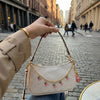 A hand holds a beige leather jewelry bag with a gold chain and pink glass charms in an outdoor city setting.