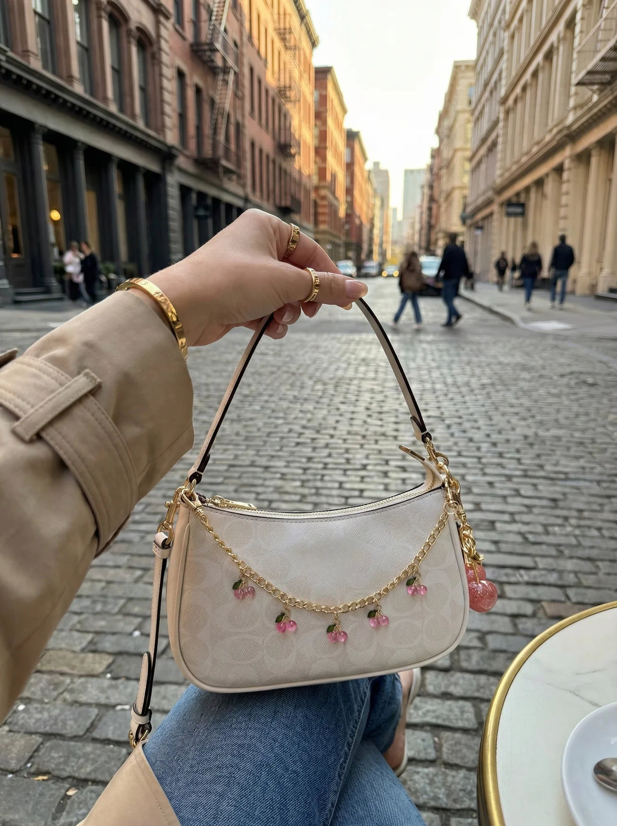 A hand holds a beige leather jewelry bag with a gold chain and pink glass charms in an outdoor city setting.