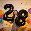 A woman holding two giant black foil balloons shaped like the number 28 in a brightly lit room.
