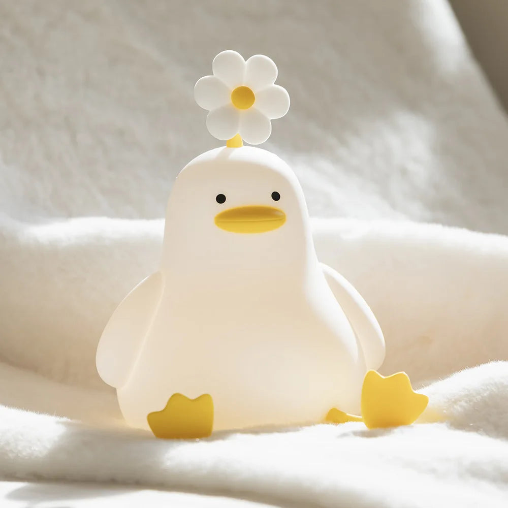 A white ceramic duck night light with a yellow flower on its head resting on a white fabric.