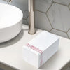 A stack of white paper dinner napkins with rose gold trim resting on a white countertop next to a modern faucet.