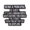 Black enamel pin with white text that reads Being a principal is easy. It's like looking both ways before you cross the stree