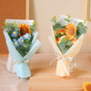 A bouquet of handcrafted crochet sunflowers and blue flowers wrapped in light blue and beige paper.