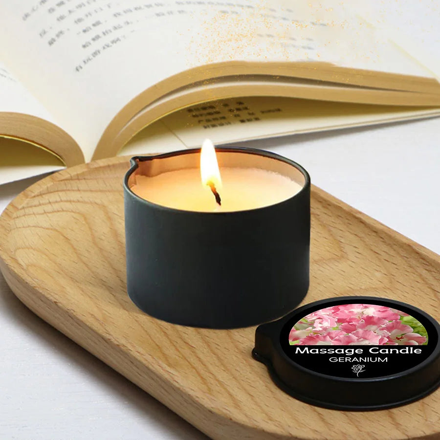 A dark candle in a holder sits on a wooden board next to the product label for an Aromatherapy Massage Candle with Geranium.