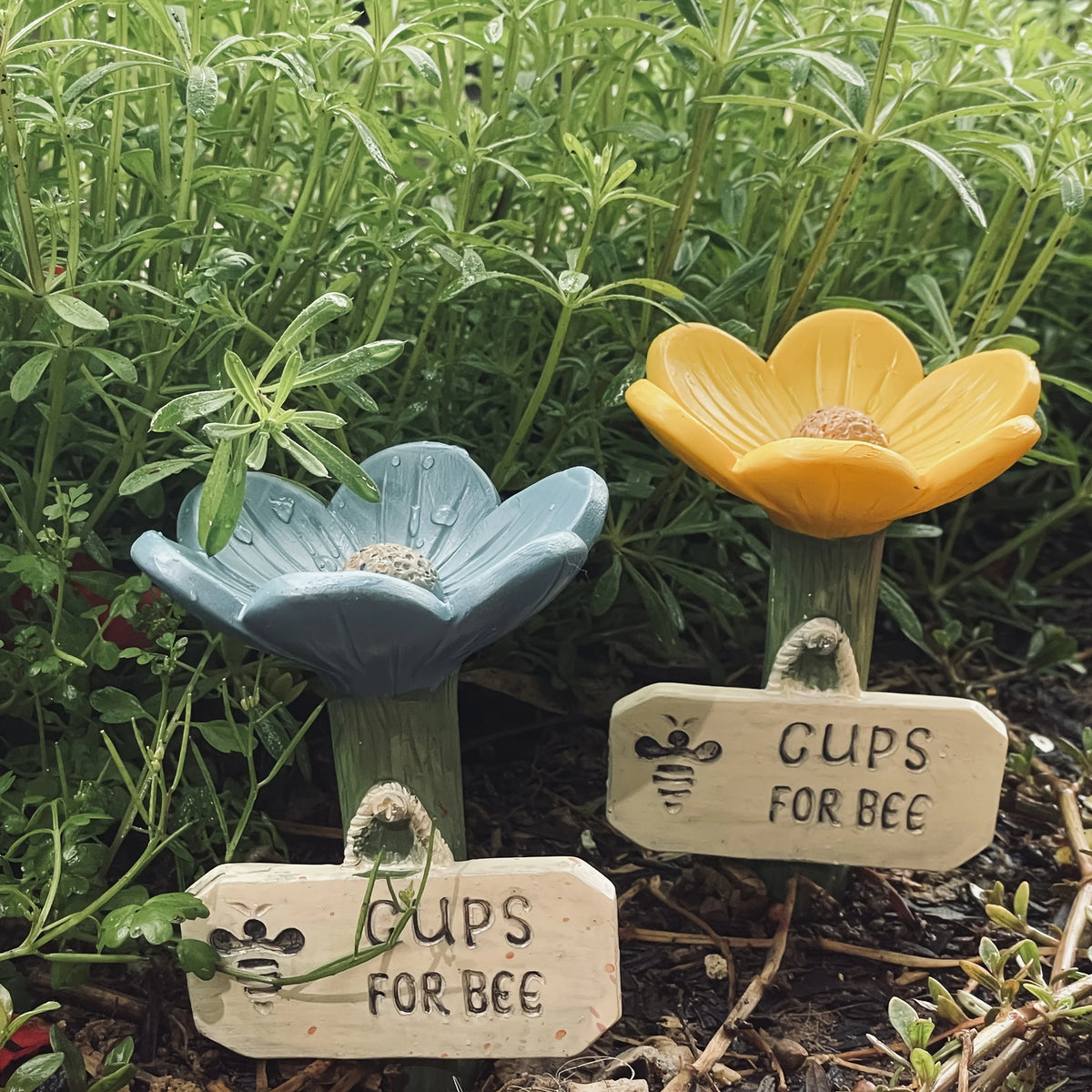 A yellow flower made of blue clay sits on a wooden stick, surrounded by green garden plants.