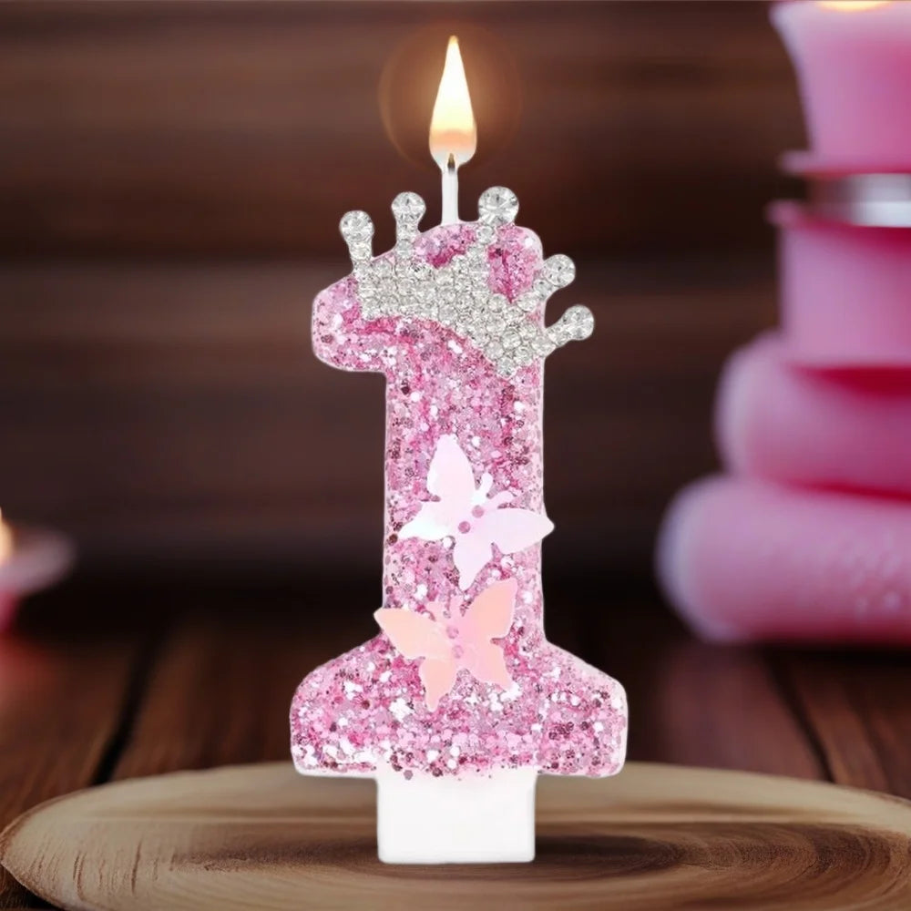 A pink glitter birthday candle shaped like the number one with a butterfly detail, lit on a wooden surface.