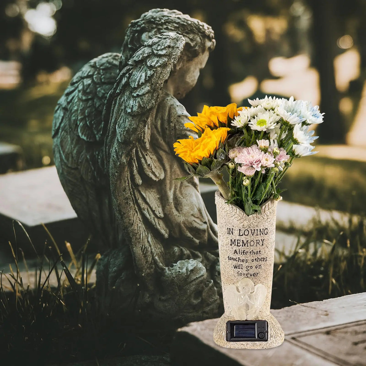 A stone angel sculpture holding a bouquet of yellow and white flowers in a solar grave memorial vase.