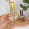 A hand holds a golden wire frame vase with thin glass tubes, set against a dark background.