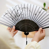A hand holding a vintage silk bamboo folding fan with black and white striped paper and delicate black line drawings of plant
