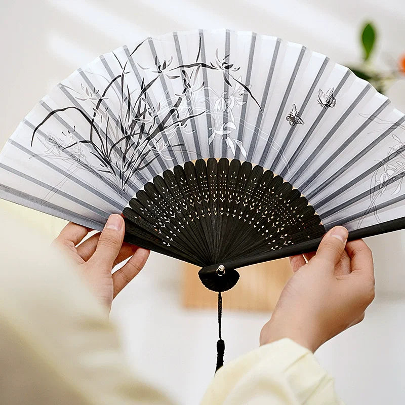 A hand holding a vintage silk bamboo folding fan with black and white striped paper and delicate black line drawings of plant
