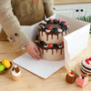 A person is preparing a cake in a white bakery box, surrounded by various pastries and ingredients.