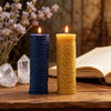 Two glowing candles in blue and yellow holders sit on a wooden surface next to a book and lavender flowers.
