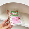 A hand holds a clear binder filled with pastel macaron-themed pages and gold hardware for K-Pop collectibles.