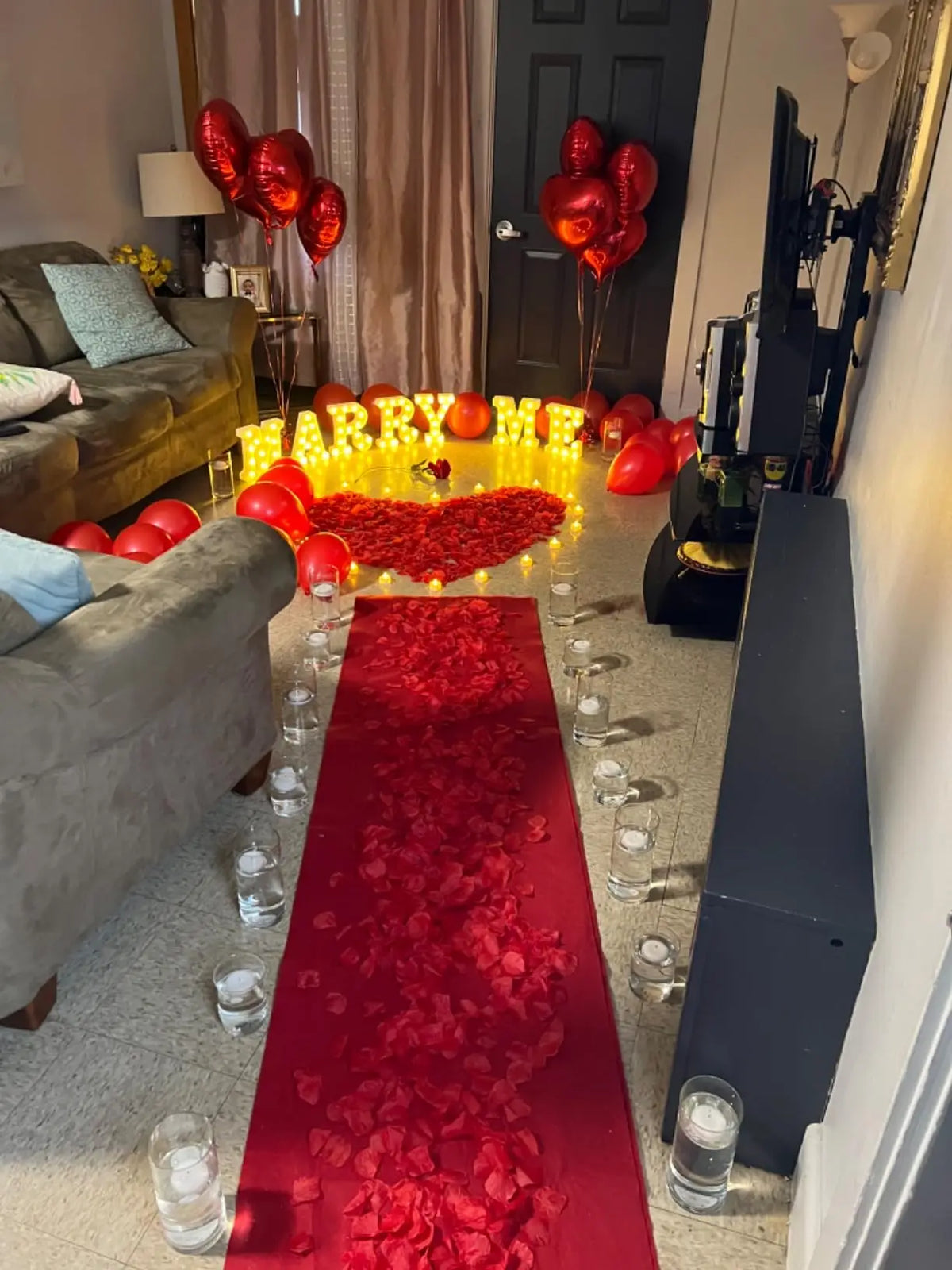 Ein Pfad aus roten Rosenblättern führt durch einen Flur mit leuchtenden 'MARRY ME'-Buchstaben und roten Ballons.