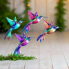 A vibrant hummingbird sculpture in iridescent pink, green, and blue perched on a green branch.