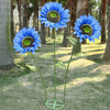 A vibrant blue silk sunflower with a yellow center is displayed on a metal base in a park setting.