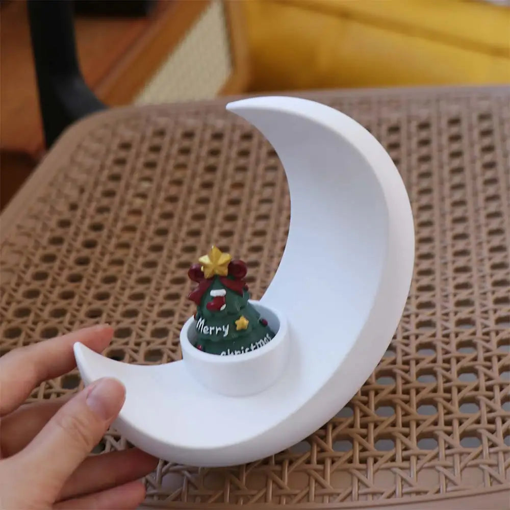 A white moon-shaped decorative vase with a green Christmas tree figurine sits on a woven surface.