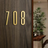 Close-up of a polished gold house number 708 mounted on dark wood paneling.