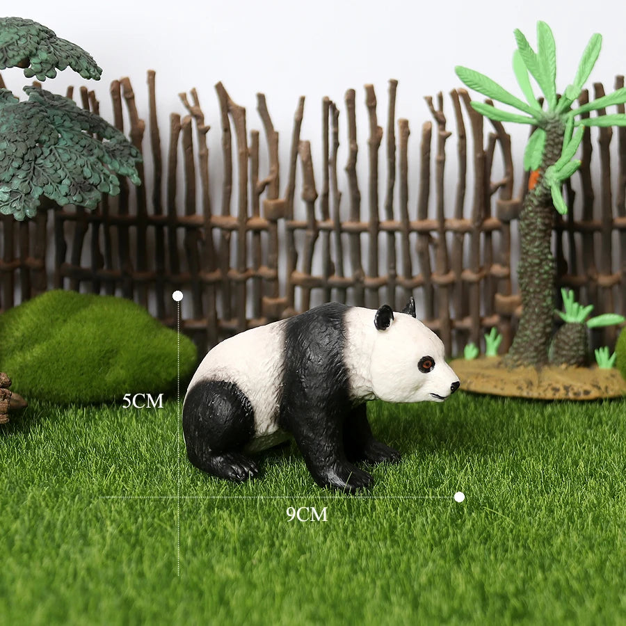 A realistic black and white panda figurine is placed on green grass in front of a wooden fence and tropical plants.