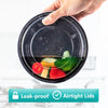 A hand is holding a round, black meal prep container with an airtight lid, showing fresh vegetables inside.
