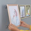 Hands are hanging a framed artwork with a light pink frame onto a gray wall using adhesive hooks.