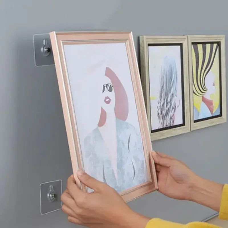 Hands are hanging a framed artwork with a light pink frame onto a gray wall using adhesive hooks.