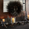 A clear glass candle holder with a versatile vase design sits on a dark wooden shelf surrounded by dried foliage and a large 