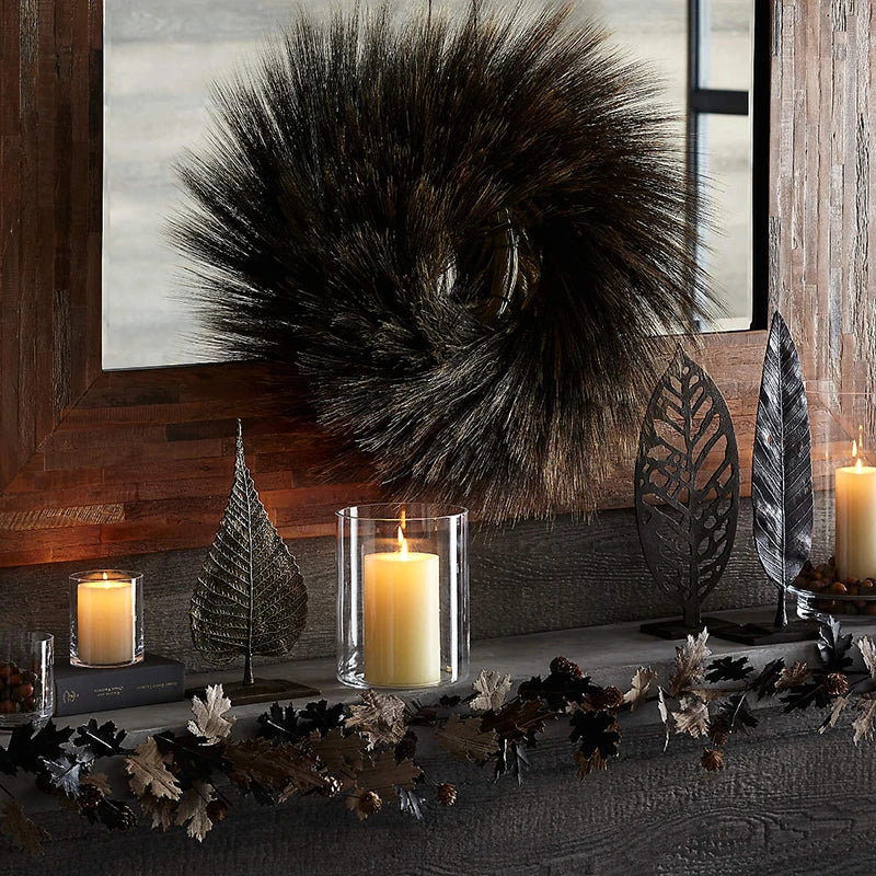 A clear glass candle holder with a versatile vase design sits on a dark wooden shelf surrounded by dried foliage and a large 