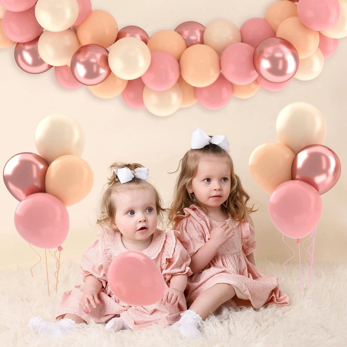 Two young girls in pink dresses sitting on a fluffy surface surrounded by pink and peach balloons