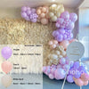 A balloon garland kit featuring purple, peach, and white balloons arranged against a fluffy beige backdrop.