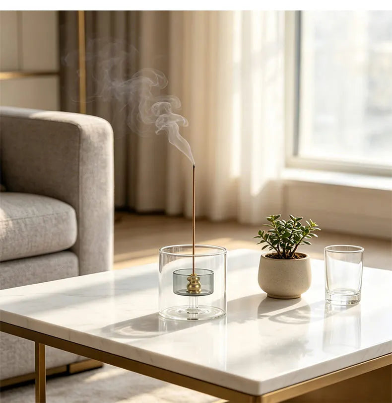 A tall, slender glass incense burner with a gourd shape and ash catcher emitting smoke on a white marble table.