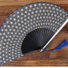 A folding hand fan with a dark patterned silk cover and a silver frame resting on a wooden surface.
