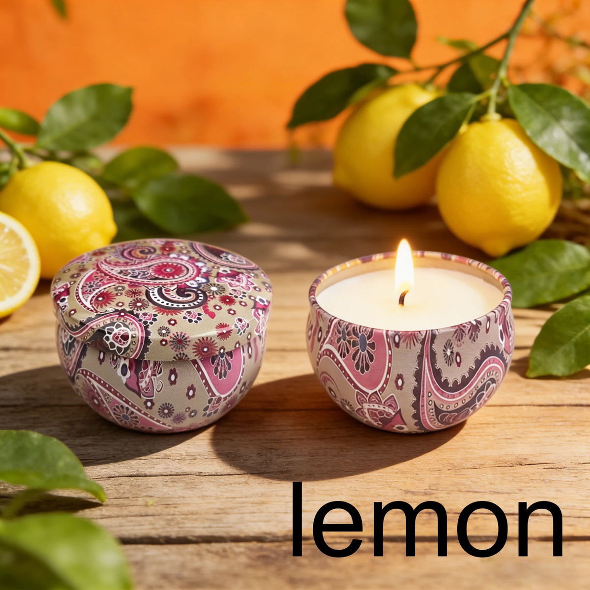 A candle in a pink patterned tin sits next to fresh lemons on a wooden surface.