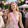 A smiling woman wearing a pink birthday crown and a sparkly pink sash at an outdoor event.