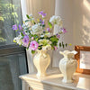 A white ceramic vase filled with purple and white flowers sits on a light-colored table next to a window.