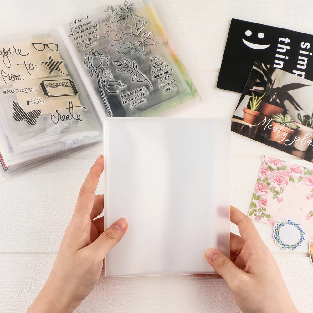 Hands hold a clear A6 storage album with pages for stickers, photos, and craft supplies next to a black card.