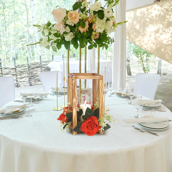 A rustic wooden wedding lantern centerpiece holder with red roses and greenery on a white tablecloth.