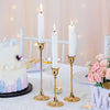 A white tiered cake decorated with purple and blue accents sits next to three tall white candles on gold stands and a bouquet