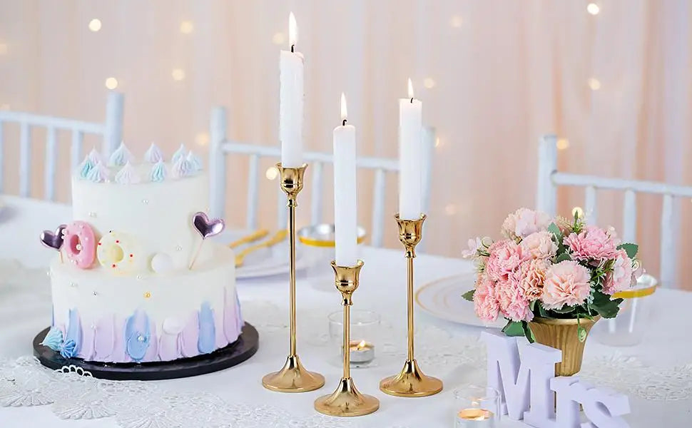A white tiered cake decorated with purple and blue accents sits next to three tall white candles on gold stands and a bouquet