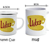 A white enamel cup and a white ceramic mug both featuring a yellow silhouette of a coffee cup with the text Luke's