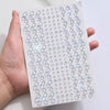 A hand holds a sheet of glitter gem stick-on facial tattoos featuring various heart and diamond shapes.